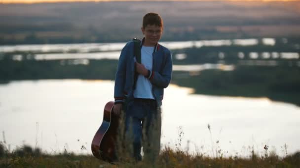 Мальчик с гитарой в синей куртке уходит с заката — стоковое видео