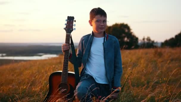 フィールドの木製ギターを立っている少年 Hes のデニム ジャケットを身に着けています サンセット — ストック動画