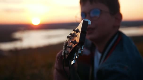 Мальчик в круглых очках играет на гитаре в поле на закате и смотрит в небо — стоковое видео