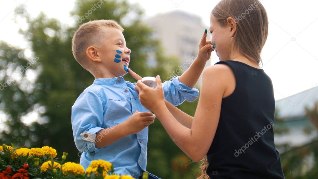 Little blonde girl and her younger friend are playing with paint in the park