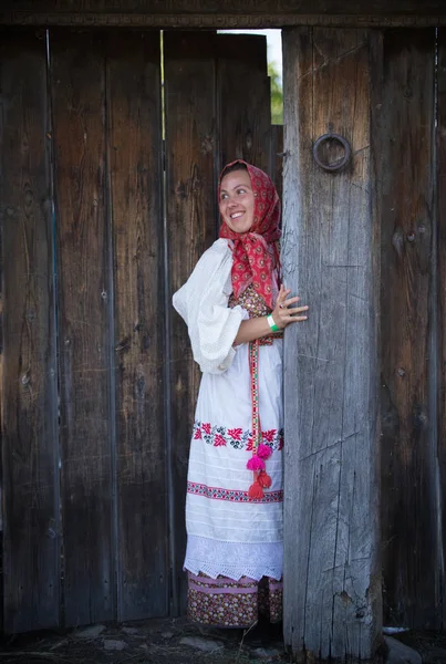 Женщина в русском народном платье и шарфе, стоящая у деревянных ворот и улыбающаяся — стоковое фото