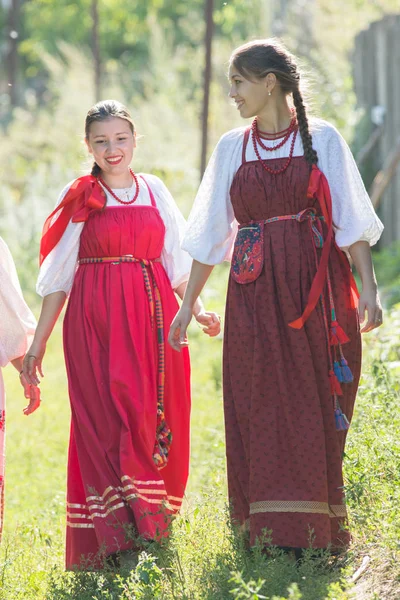 Duas meninas bonitas jovens em vestidos nacionais russos estão andando na grama e sorrindo — Fotografia de Stock