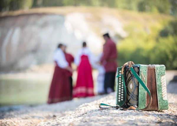 Aufnahme des Akkordeons, vor dem Hintergrund einer Gruppe junger Leute in roten russischen Nationaltrachten — Stockfoto