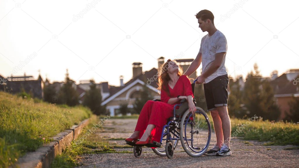 A beautiful girl with red hair in a red dress is sitting in a wheelchair and smiling at her boyfriend who is standing next to her, behind her