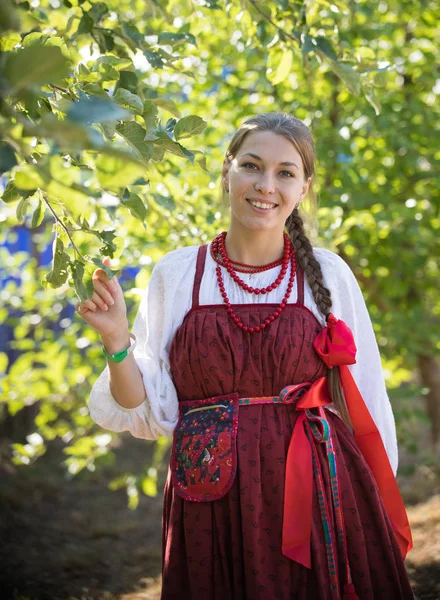 Smiling young woman in russian folk costume on the foliage background — Stock Photo, Image