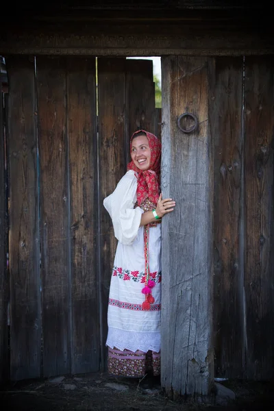 A woman in a Russian folk dress and a scarf standing at the wooden gate — Stock Photo, Image