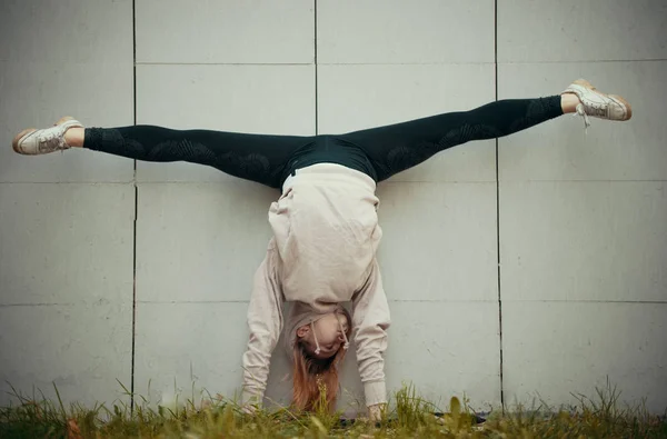 Acrobat meisje in een beige hoodie voert de splitsingen, terwijl staande op op een achtergrond van de muur handen. Brede shot — Stockfoto