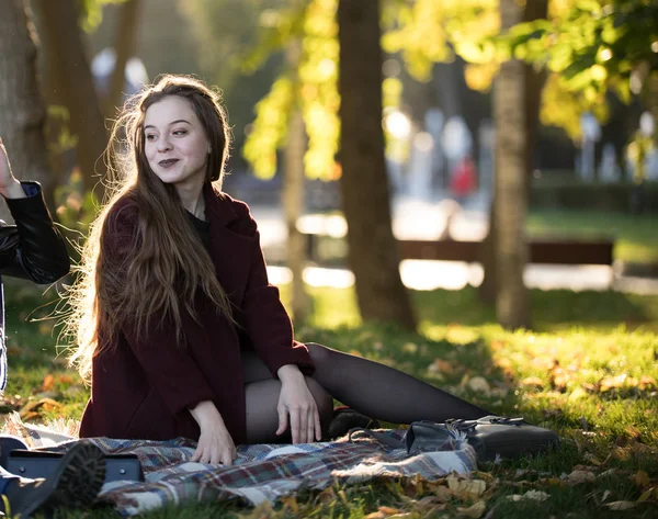 Дівчина в бургундському пальто, що сидить на плед в осінньому сонячному парку — стокове фото
