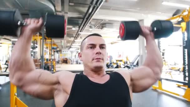 Bodybuilder dans un débardeur noir effectue un exercice de presse épaule haltère assis dans une chaise — Video