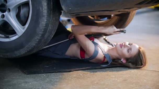 Sexy chica mecánica acostada debajo del coche y alcanzando la llave, sonriendo — Vídeos de Stock