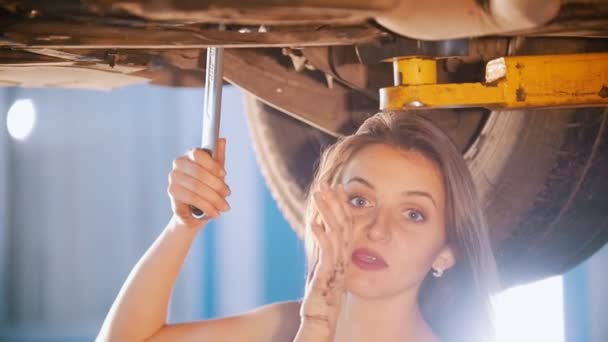 Σέξι κορίτσι μηχανικός κάτω από το αυτοκίνητο με ένα κλειδί, μιλώντας σε οπερατέρ και χαμόγελο — Αρχείο Βίντεο