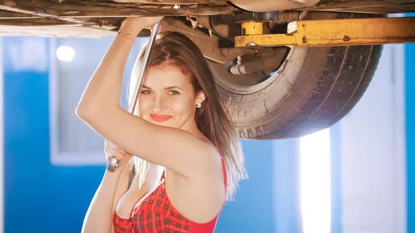 Sexy mechanic meisje onder de auto met een spanner, glimlachend — Stockfoto