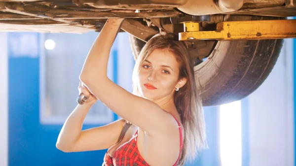 Sexy mechanic meisje onder de auto met een spanner, kijken in de camera — Stockfoto