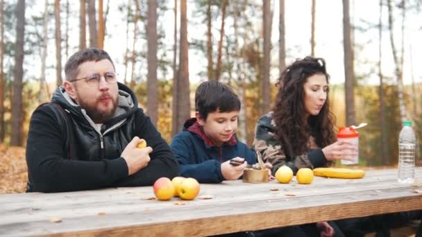 Mamá, papá y el niño sentados uno al aire libre en el bosque en la mesa y disfrutar de la comida . — Vídeo de stock