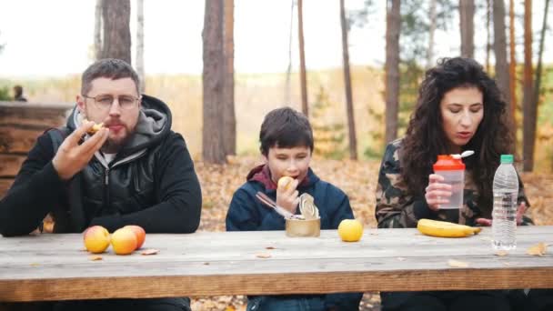 Mama, Tata i dziecko siedzi w pobliżu do siebie na zewnątrz w lesie przy stole i zjeść posiłek. Widok z przodu — Wideo stockowe