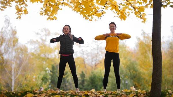 Meninas acromáticas em jaquetas aquecendo do lado de fora antes de treinar no parque. Mãos para os lados — Fotografia de Stock