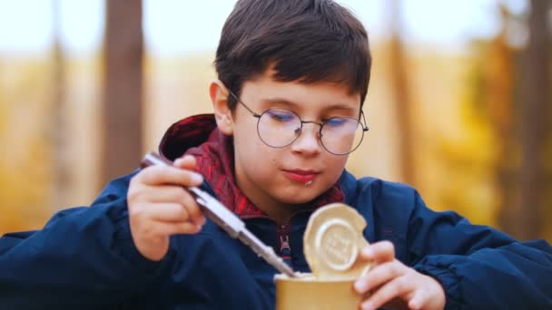 Un niño en vasos redondos comiendo carne estofada enlatada usando un cuchillo — Vídeos de Stock