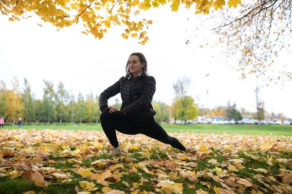 Outono foto de desportivo Jovem mulher de casaco preto que se estende na floresta de manhã contra o fundo das árvores — Fotografia de Stock