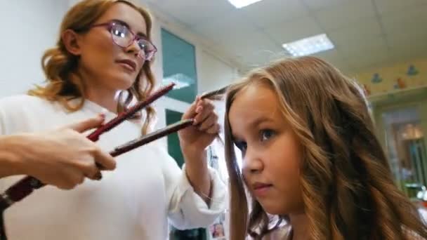 Stylist curling långt hår för lilla söta tjejen, använder elektriska locktång. Närbild — Stockvideo