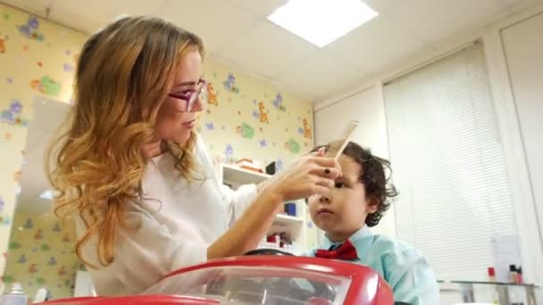 Kinderen barbershop. Kleine jongen zitten in speelgoedauto en stylist kamt zijn haar — Stockvideo