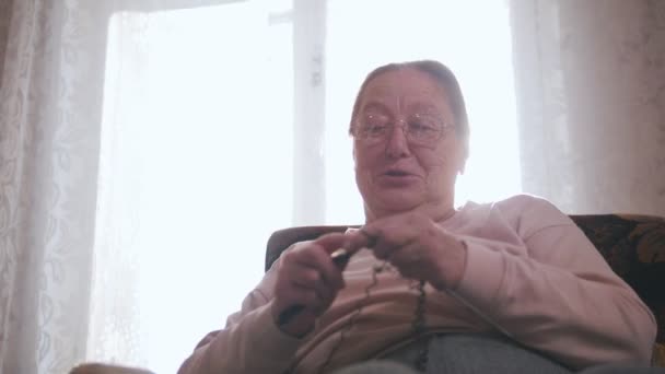 An elderly woman sits in a chair and knits on the background of the window — Stock Video