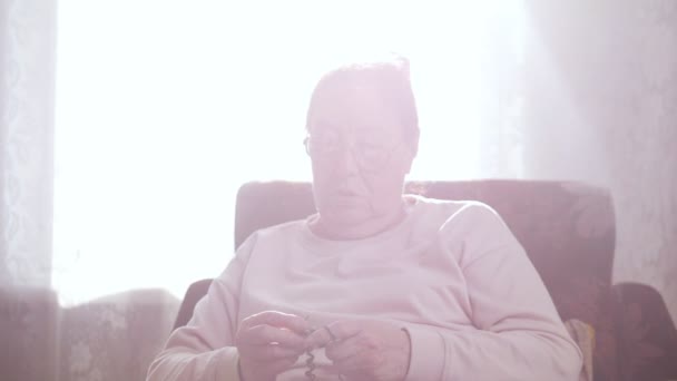 An elderly woman enjoys sitting in a chair and knitting on the background of the window — Stock Video