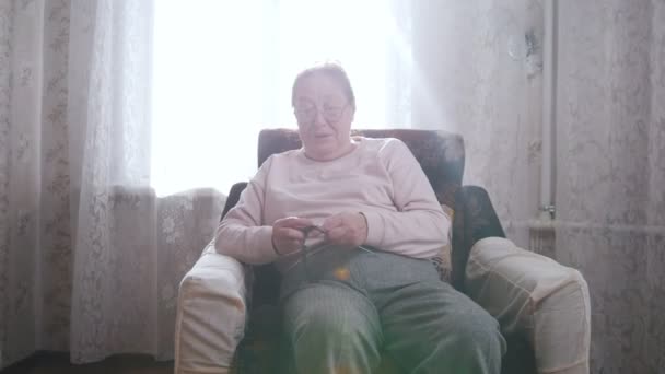 Starší žena se těší její koníček, sedí v křesle a pletení na pozadí okna. — Stock video