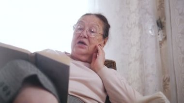 Yaşlı bir kadın bir sandalyede oturuyor ve yüksek sesle pencerenin arka plan üzerinde okuma seviyor. Yakın çekim