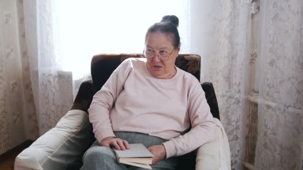 Starší žena se těší sedí v křesle a čte nahlas na pozadí okna. — Stock video