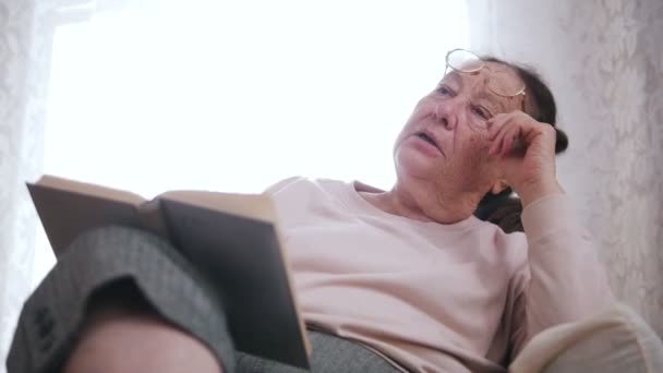 Eine ältere Frau sitzt auf einem Stuhl und liest ein Buch auf dem Fensterhintergrund. — Stockvideo