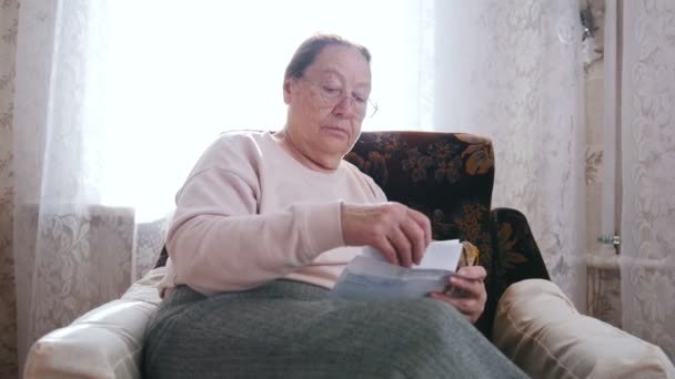 Eine ältere Frau sitzt auf einem Stuhl und öffnet einen Brief auf dem Hintergrund des Fensters. — Stockvideo