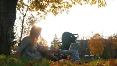 Genç çimde oturma ve küçük bebek oğlu ile oynarken anne. Sonbahar yaprakları ile oynamak