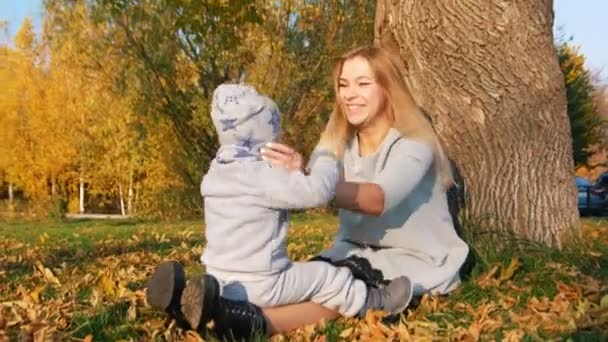 Νεαρή μητέρα κάθεται στο γρασίδι με το μικρό μωρό, χαμογελώντας. Κουνώντας το χέρι του στον κάμεραμαν. — Αρχείο Βίντεο