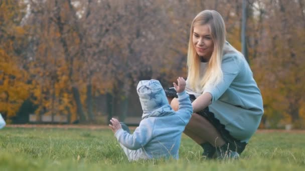 Young mother with a baby walking in the autumn park. Baby lies on the grass, mother helps him to get up — Stock Video