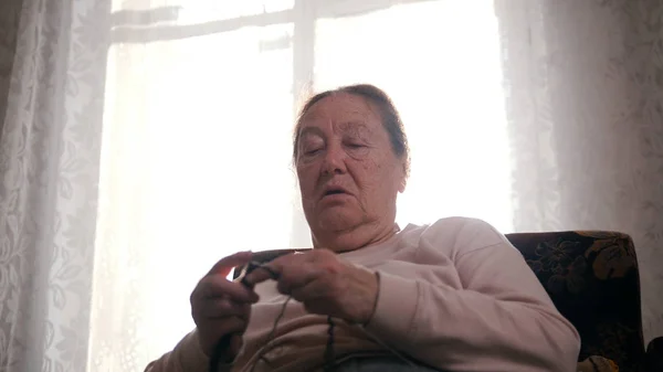 En äldre kvinna som sitter i en stol och stickat på bakgrunden av fönstret. Närbild — Stockfoto
