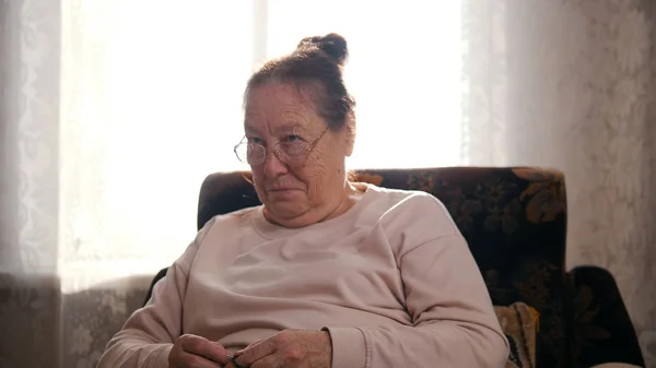 Bir sandalyede oturan gözlüklü yaşlı bir kadın ve pencerenin arka plan üzerinde dokuma kumaşlar — Stok fotoğraf