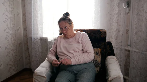 Una anciana con su pasatiempo sentada en una silla y tejiendo en el fondo de la ventana, mirando enojada — Foto de Stock