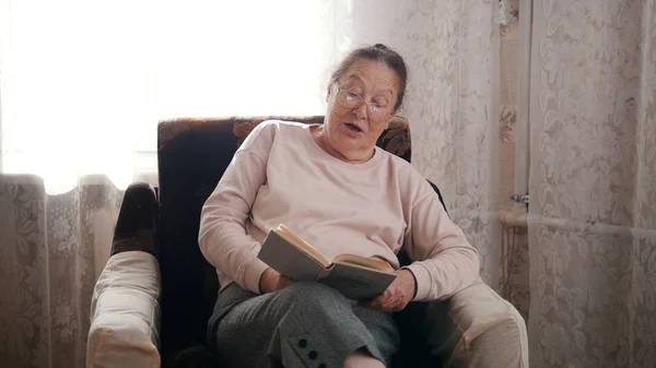 Starsza kobieta siedzi na krześle i czytanie na głos, na tle okna. — Zdjęcie stockowe