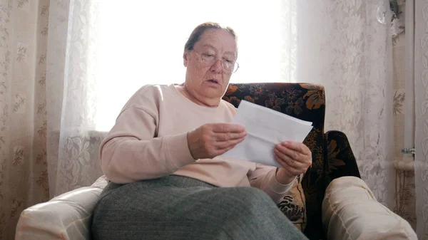 Пожилая женщина, сидящая в кресле и читающая что-то, выглядящая удивленной не в хорошем смысле . — стоковое фото