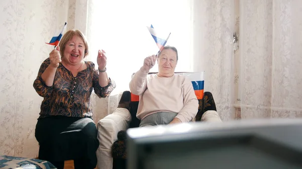 Две пожилые женщины смотрят телевизор и размахивают российскими флагами — стоковое фото