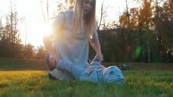 Madre joven con su bebé jugando en el parque de otoño al atardecer. Bebé se acuesta en la hierba . — Vídeos de Stock