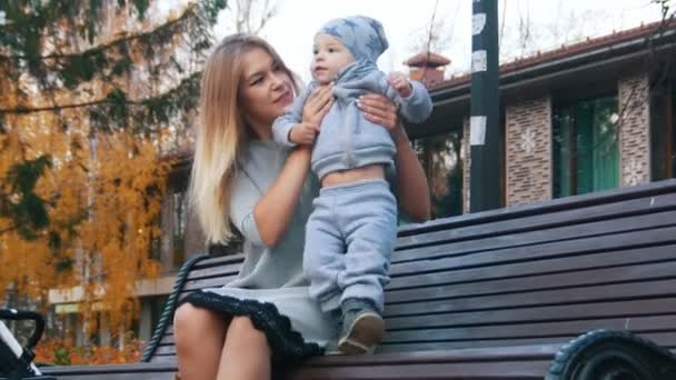 Jonge moeder in jurk met haar kleine baby zittend op het bankje in herfst park en spelen. — Stockvideo