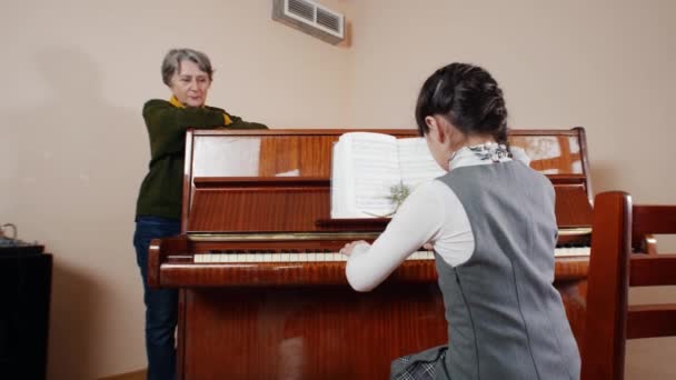 Урок музыки. Девушка играет на пианино, старший учитель стоит рядом с пианино. Слайдерный вид на игру со спины девочки — стоковое видео