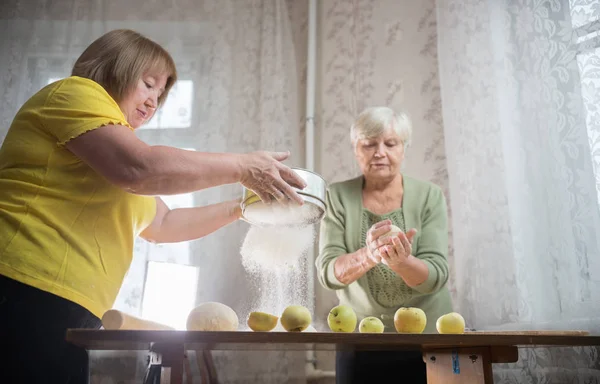 Две пожилые женщины делают яблочные пирожки дома. Просеивание муки на столе — стоковое фото