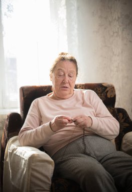 Yaşlı bir kadın onun hobi bir koltukta oturan ve pencerenin arka plan üzerinde örgü sahiptir.