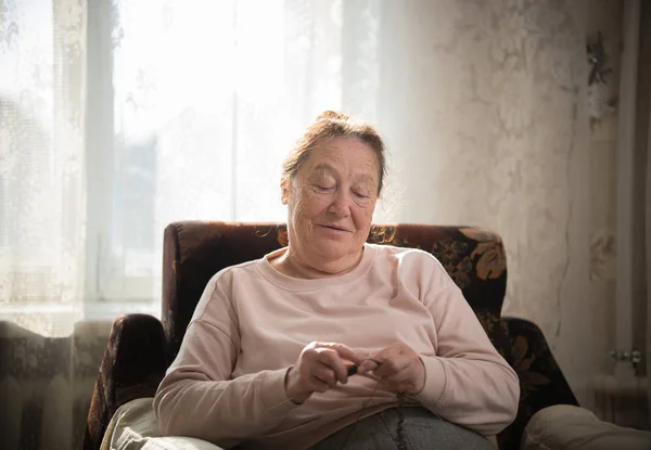 Une femme caucasienne âgée jouit de son passe-temps assis dans un fauteuil et tricote sur le fond de la fenêtre . — Photo