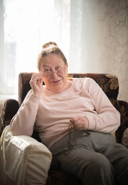 Une femme caucasienne âgée en lunettes jouit de son passe-temps assis dans un fauteuil et tricotant sur le fond de la fenêtre . — Photo