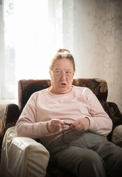 Hobby. An elderly woman enjoys her hobby sitting in a armchair and knitting on the background of the window. — Stock Photo, Image