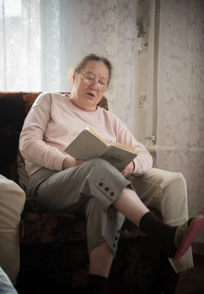 Una anciana mujer caucásica en gafas disfruta de su hobby sentada en un sillón y leyendo libros en el fondo de la ventana . — Foto de Stock