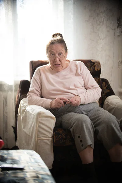 Les passe temps. Une femme âgée en lunettes jouit de son passe-temps assis dans un fauteuil et tricote sur le fond de la fenêtre . — Photo